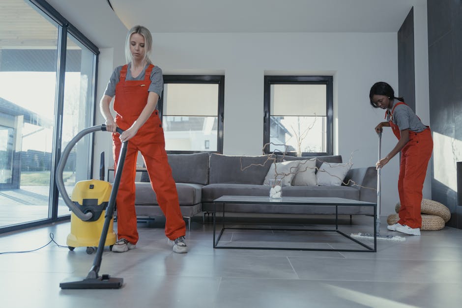 Two professional cleaners wearing orange uniforms and grey shirts are engaged in surface cleaning within a modern living room. The cleaner on the left is using a yellow vacuum cleaner to clean the lig