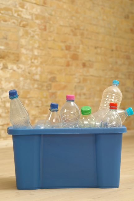 Close-up image of a clear plastic recycling bin with a green recycling symbol sticker on the front, placed on a light-colored tiled floor. Behind the bin, there is a partially visible black dishwasher