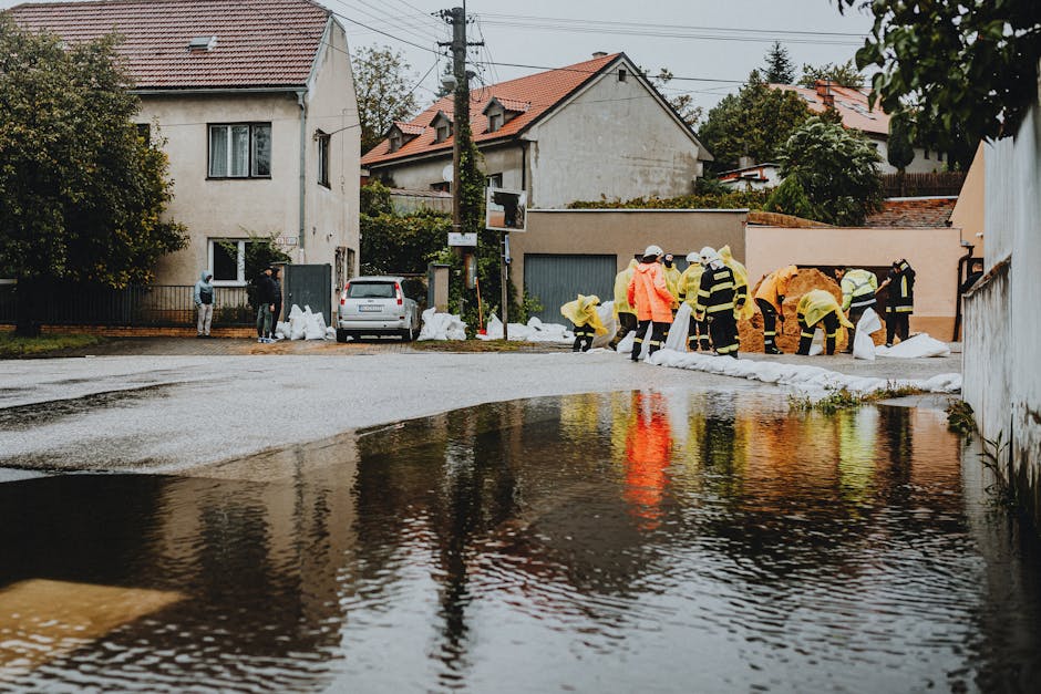 Aerial view of a residential neighborhood with numerous houses, some of which are partially submerged due to flooding. The flooded streets and properties show water covering driveways and yards, with 