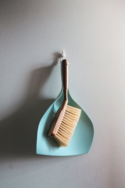 A close-up of a light blue dustpan hanging on a plain, light grey wall, with a wooden-handled scrub brush resting inside it. The brush has beige bristles and is positioned with the handle leaning to the left, partially overlapping the dustpan’s edge. The scene is illuminated by soft, natural light, emphasizing the cleanliness and tidy arrangement, typical of surface cleaning and maintenance in a domestic or commercial setting. This image reflects the importance of detailed cleaning tools for effective sanitisation, as promoted by Kentish Town Cleaners' end of tenancy cleaning checklist for landlords, available at kentishtown-cleaners.co.uk.